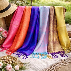 Puede incluir: Una exhibición de coloridas bufandas en tonos rosa, naranja, azul, morado y dorado. Las bufandas presentan patrones intrincados y detalles de flecos. La imagen también incluye un sombrero de paja y una cesta de flores, sugiriendo un tema veraniego.