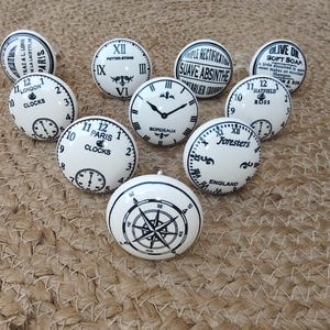 Peut inclure: Ensemble de boutons de meubles en céramique blanche avec des motifs noirs. Les boutons présentent des cadrans d'horloge, des roses des vents et des textes tels que "London", "Paris" et "Bordeaux". Disposition sur une surface tissée beige.