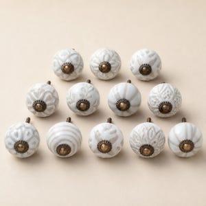 May include: A collection of twelve round ceramic cabinet knobs arranged in three rows. Each knob features a white base with light brown or beige embossed patterns, including floral, striped, and geometric designs, finished with a brass centre.