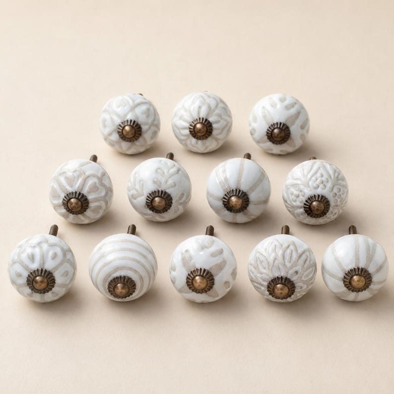 May include: A collection of twelve round ceramic cabinet knobs arranged in three rows. Each knob features a white base with light brown or beige embossed patterns, including floral, striped, and geometric designs, finished with a brass centre.