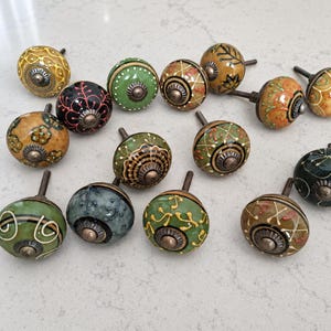 Può includere: Una collezione di tredici pomelli rotondi in ceramica per mobili con vari motivi dipinti a mano in tonalità di verde, giallo, arancione, nero e blu. Ogni pomello ha un bullone metallico centrale e una base decorativa in metallo scuro.