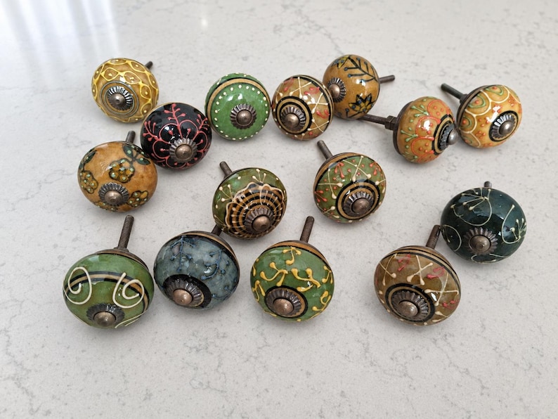 May include: A collection of thirteen round ceramic cabinet knobs featuring various hand-painted patterns in shades of green, yellow, orange, black, and blue. Each knob has a central metallic bolt and a decorative metal base with a dark finish.