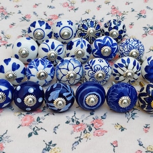 Może przedstawiać: Kolekcja dekoracyjnych ceramicznych gałek do szafek w różnych niebiesko-białych wzorach kwiatowych i geometrycznych. Każda gałka ma srebrną metalową podstawę. Gałki ułożone są na tkaninie we wzory kwiatowe.
