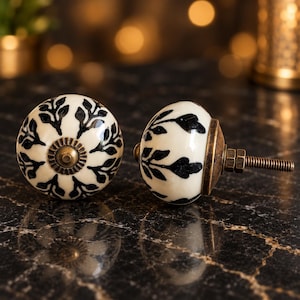 May include: Two round cabinet knobs featuring a cream-colored ceramic surface with a black leaf pattern. Each knob has a central antique brass-toned metal accent and a threaded metal bolt extending from the back for installation purposes.