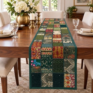 Könnte beinhalten: Ein rechteckiger Tischläufer mit einem Patchwork-Design aus verschiedenen Stoffquadraten in Grün-, Gold-, Rot- und dunklen Petroltönen. Der Läufer zeigt vielfältige Texturen, florale Muster und metallische Akzente, eingefasst von einem dunkelgrünen Rand.