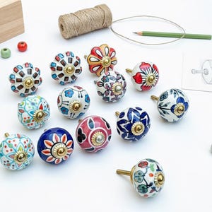 Può includere: Una collezione di pomelli decorativi in ceramica con vari colori e motivi floreali. Ogni pomello presenta una base in ottone e un design unico dipinto a mano, inclusi motivi floreali blu, rossi e bianchi. Una matita, dello spago e un cassetto di legno sono anche nell'immagine.