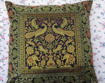 May include: Square decorative pillow with a detailed pattern featuring peacocks, elephants, and floral motifs in shades of gold, green, and orange on a black background. The pillow is placed on a floral patterned bedspread.