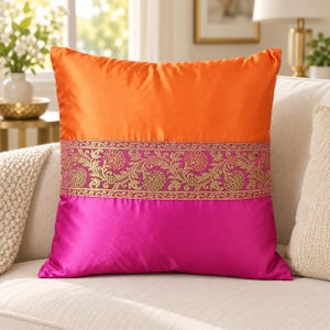 Puede incluir: Un cojín cuadrado con un diseño horizontal de bloques de color, con una sección superior naranja vibrante y una sección inferior fucsia brillante, separadas por una ancha banda central de intrincado bordado floral dorado sobre fondo rosa.