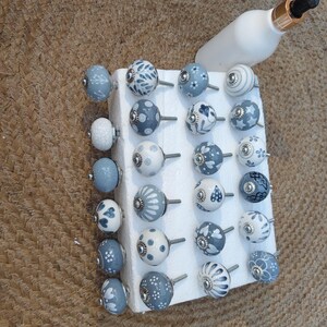 Op de afbeelding: Een verzameling decoratieve keramische kastknoppen in verschillende ontwerpen en tinten blauw en wit. De knoppen worden getoond op een wit schuimblok, met een witte fles met een gouden en zwarte dop op de achtergrond.