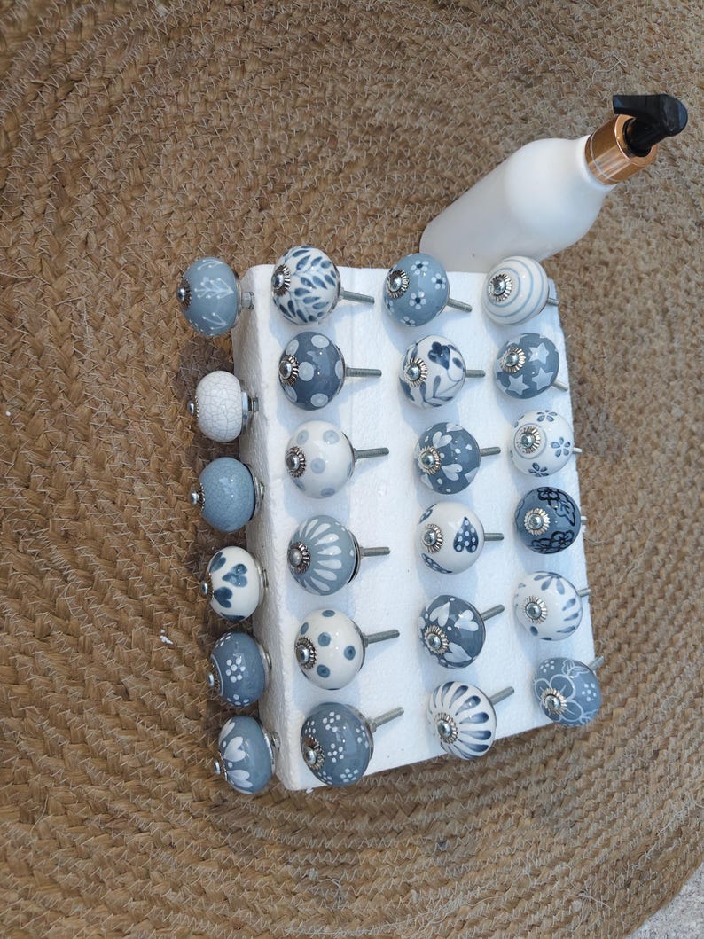 Op de afbeelding: Een verzameling decoratieve keramische kastknoppen in verschillende ontwerpen en tinten blauw en wit. De knoppen worden getoond op een wit schuimblok, met een witte fles met een gouden en zwarte dop op de achtergrond.