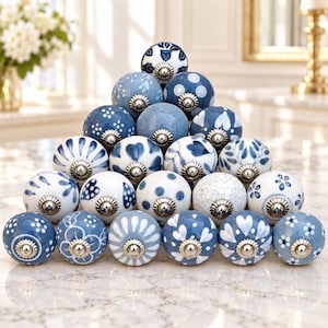 May include: A collection of twenty-one round ceramic cabinet knobs arranged in a pyramid. Each knob features a unique blue and white pattern, including floral, polka dot, heart, and crackle designs, finished with a metallic silver-toned base.