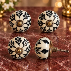 May include: Four round ceramic cabinet knobs featuring a cream-colored base with a black leaf pattern radiating from the center. Each knob has a central brass-toned metal accent and a threaded metal bolt extending from the back for installation.