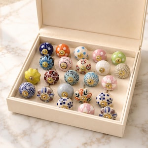 Pode incluir: Uma cole&ccedil;&atilde;o de vinte e um puxadores de cer&acirc;mica decorativos para arm&aacute;rios dispostos numa caixa de armazenamento cor de creme. Cada puxador apresenta um padr&atilde;o &uacute;nico, incluindo bolinhas, motivos florais e desenhos geom&eacute;tricos em v&aacute;rias cores com ferragens douradas.