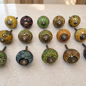 May include: A collection of fourteen round ceramic cabinet knobs arranged in three rows. Each knob features a unique hand-painted pattern in various colours including green, yellow, black, and orange, with a central metallic screw and base.