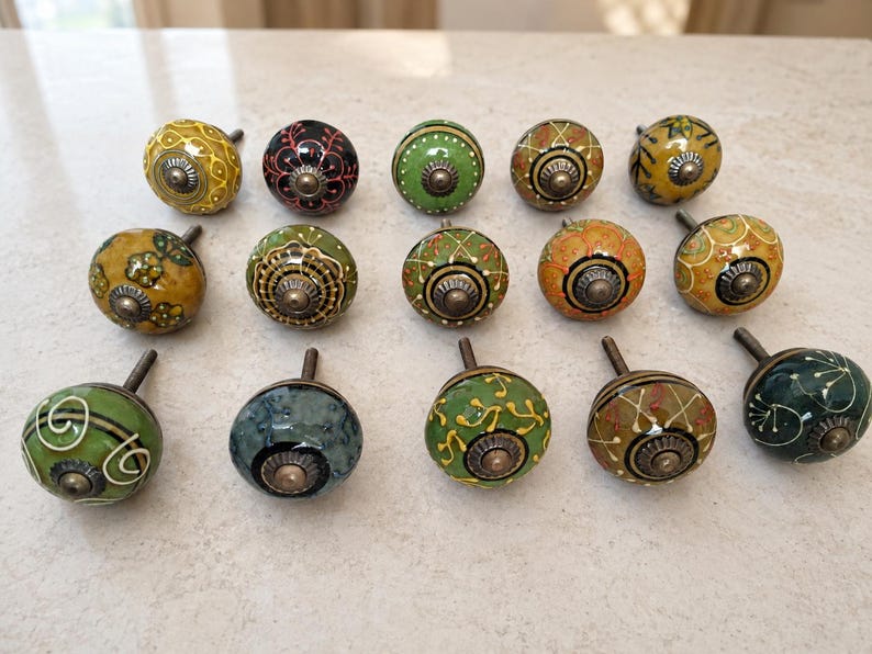 May include: A collection of fourteen round ceramic cabinet knobs arranged in three rows. Each knob features a unique hand-painted pattern in various colours including green, yellow, black, and orange, with a central metallic screw and base.