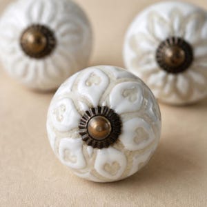 May include: Three round ceramic cabinet knobs featuring a white floral pattern with heart-shaped petals over a beige base. Each knob has a central dark bronze-toned metal hardware piece with a ridged edge and a small, smooth, rounded centre point.