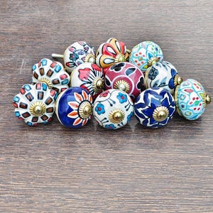 Pu&ograve; includere: Una collezione di pomelli decorativi in ceramica con vari motivi floreali e geometrici colorati. Ogni pomello presenta un design unico con una base in metallo color ottone. I pomelli sono disposti su una superficie di legno.