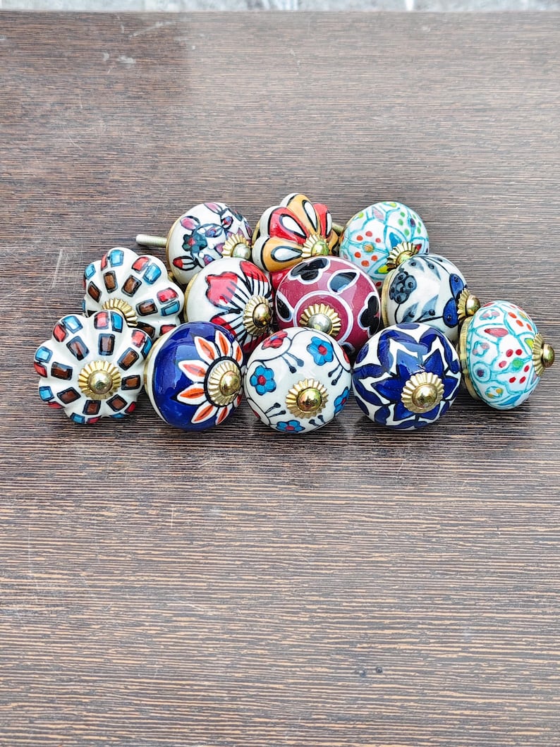 Pu&ograve; includere: Una collezione di pomelli decorativi in ceramica con vari motivi floreali e geometrici colorati. Ogni pomello presenta un design unico con una base in metallo color ottone. I pomelli sono disposti su una superficie di legno.