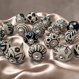 Pode incluir: Uma coleção de puxadores redondos de cerâmica para armários com vários padrões pintados à mão em preto e branco, incluindo designs florais, geométricos e listrados. Cada puxador tem um acessório central de metal prateado.
