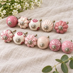 Op de afbeelding: Een verzameling van tien decoratieve kastknoppen in diverse tinten roze, crème en wit. De knoppen hebben uiteenlopende patronen zoals bloemenprints, strepen, stippen en geschulpte randen, elk afgewerkt met een centrale metalen schroef.