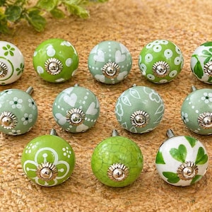 May include: A collection of eleven decorative ceramic knobs in shades of green and white. Each knob features a unique floral or heart design, with a silver-colored metal base. The knobs are arranged on a textured, light brown surface.