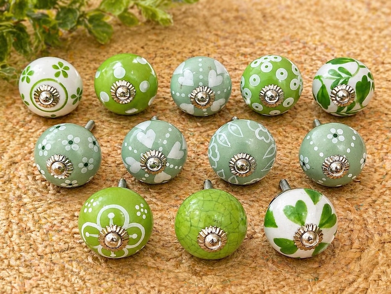 Pode incluir: Uma cole&ccedil;&atilde;o de onze puxadores decorativos de cer&acirc;mica em tons de verde e branco. Cada puxador apresenta um design floral ou de cora&ccedil;&atilde;o &uacute;nico, com uma base de metal prateado. Os puxadores est&atilde;o dispostos sobre uma superf&iacute;cie texturizada castanho-claro.