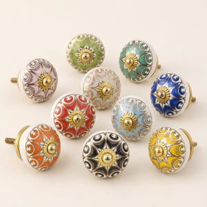Pu&ograve; includere: Una collezione di nove pomelli rotondi in ceramica per mobili, ognuno caratterizzato da un motivo floreale multicolore unico con accenti dorati e un tappo centrale in metallo dorato. I pomelli sono disposti in gruppo, mostrando colori tra cui rosso, blu, verde, giallo, arancione, rosa, nero e bianco.