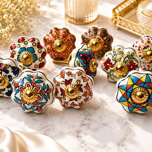 May include: A collection of ten ceramic drawer knobs featuring various colourful floral and geometric patterns. Each knob has a scalloped, flower-like shape and a central gold-toned metal hardware piece. The designs include vibrant red, blue, yellow, and green motifs painted on white ceramic surfaces.