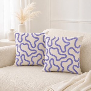 May include: Two square throw pillows featuring a cream-colored base fabric with a raised, textured purple tufted pattern consisting of irregular, winding, and interconnected organic shapes that cover the entire front surface of each pillow.