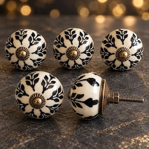May include: Five round ceramic cabinet knobs featuring a cream-colored base with a black leaf pattern. Each knob has a central antique brass-toned metal accent and a threaded metal bolt extending from the back for installation purposes.