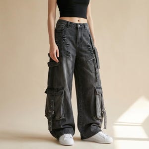 May include: High-waisted charcoal grey denim cargo trousers featuring a wide-leg silhouette, distressed detailing on the thighs, multiple large side pockets with buckle straps, and a zippered pocket on the left leg, paired with white trainers.