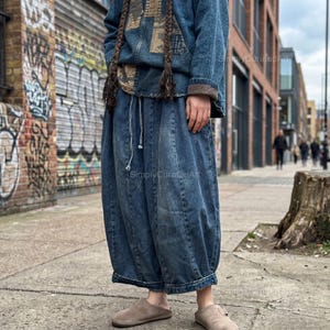 Peut inclure: Une personne porte une veste en jean à capuche et taille à cordon, assortie d'un pantalon en jean. Le pantalon a une forme volumineuse unique. La tenue est complétée par des chaussures à enfiler en daim. Le fond présente un mur de briques avec des graffitis.