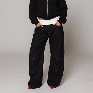 May include: Dark wash denim jeans with a wide leg design. The jeans feature decorative stitching in a contrasting color along the seams. The model is wearing a black zip-up hoodie and red shoes. The waist is accentuated with a white band.