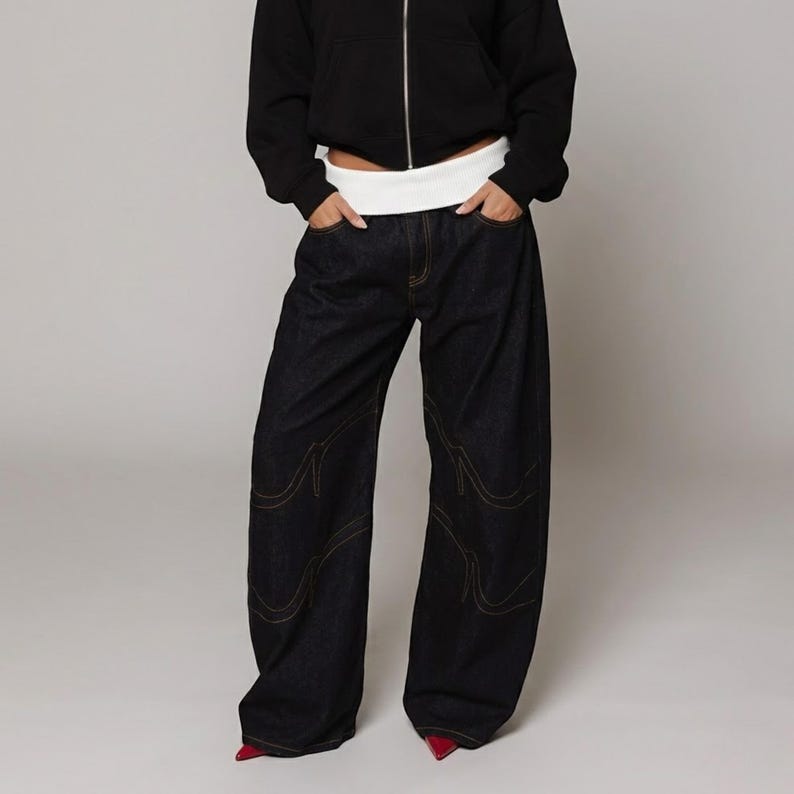 May include: Dark wash denim jeans with a wide leg design. The jeans feature decorative stitching in a contrasting color along the seams. The model is wearing a black zip-up hoodie and red shoes. The waist is accentuated with a white band.