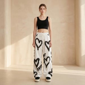 May include: A black sleeveless crop top paired with white wide-leg trousers featuring a black spray-painted heart pattern and drawstring waist. The trousers include black text graphics integrated with the heart designs and are worn with black shoes.