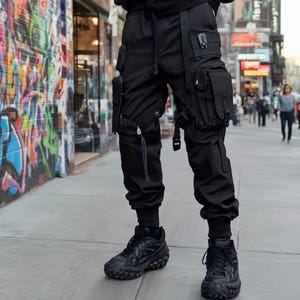 May include: Black cargo trousers with multiple pockets and straps, worn with black trainers. The trousers have a modern, urban design, featuring various compartments and adjustable elements. The trainers have a textured sole.