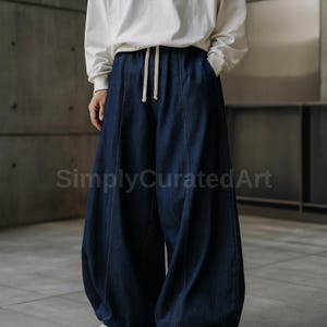 Könnte beinhalten: Dunkelblaue Jeanshose mit weitem, lockerem Schnitt. Die Hose hat einen Kordelzug in der Taille und Taschen. Der Text "SimplyCuratedArt" ist unten links im Bild sichtbar. Das Model trägt ein weißes Langarmshirt.