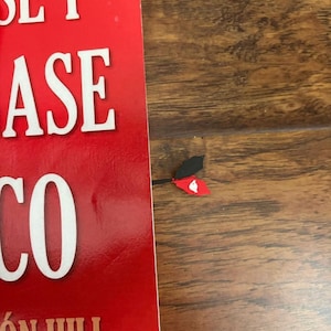 May include: A red book with white text and a decorative bookmark. The bookmark has a black and red design with a white heart shape. The book's cover is red with white text.