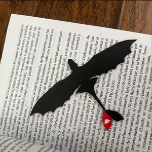 May include: Black paper cut-out of a dragon-like creature with a red heart-shaped flag, resting on the pages of a book. The dragon is silhouetted against the text, creating a striking contrast. A unique bookmark.