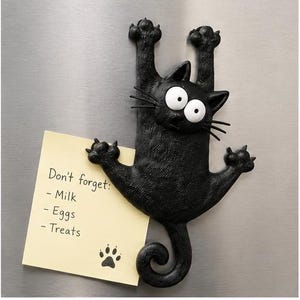 May include: A black cat-shaped refrigerator magnet with wide white eyes and a surprised expression, holding a square yellow sticky note that reads: Don't forget: - Milk - Eggs - Treats, with a small black paw print icon at the bottom.