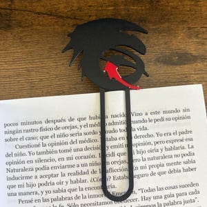 May include: Black metal bookmark featuring a stylized dragon design with a red arrow accent. The bookmark is inserted into a book with Spanish text. A unique accessory for book lovers and fans of fantasy.