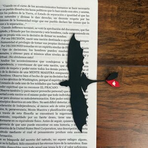 May include: Black dragon-shaped bookmark with red and white accents, placed on a book page. The bookmark is a silhouette, with a detailed wing and tail design. The book is open, with text visible.