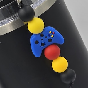 Könnte beinhalten: Ein schwarzer Becher mit einem Perlenanhänger. Der Anhänger zeigt einen blauen Videospiel-Controller, rote und gelbe Perlen sowie schwarze Perlen. Der Anhänger ist an einem weißen Kunststoffring befestigt.