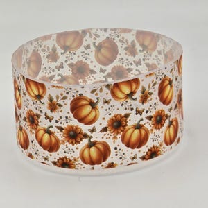 May include: A cylindrical, translucent resin ring featuring a repeating pattern of orange pumpkins, brown sunflowers, small butterflies, and delicate foliage. The design is printed on the clear material, creating a layered visual effect.