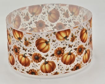 Puede incluir: Un anillo cilíndrico de resina translúcida con un patrón repetitivo de calabazas naranjas, girasoles marrones, pequeñas mariposas y follaje delicado. El diseño está impreso sobre el material transparente, creando un efecto visual en capas.