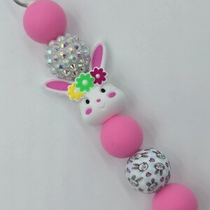 May include: A pink and white beaded keychain featuring a white bunny with a flower crown. The keychain includes pink and crystal-like beads, and a bead with a bunny and heart pattern. A silver clasp is at the top.