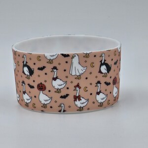 May include: A cylindrical ceramic bowl with a light peach background featuring a repeating pattern of white geese dressed in various Halloween costumes, including ghosts, pumpkins, and capes, interspersed with small black bats and yellow stars.