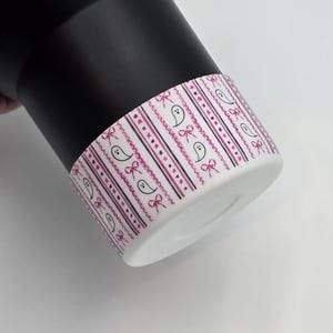 May include: Black tumbler with a white base featuring a repeating pattern of pink bows, stars, and ghost-shaped figures. The design is set against a striped background. The tumbler is designed for beverages.