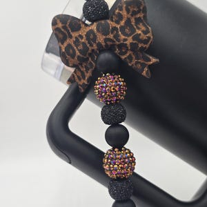 Puede incluir: Un vaso negro con un dije decorativo. El dije presenta un lazo con estampado de leopardo, cuentas negras y cuentas de cristal iridiscentes. El dije está sujeto al asa del vaso.