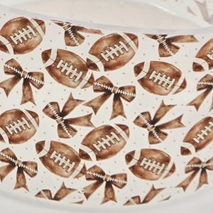 May include: A white background is covered with a pattern of brown footballs and bows. The footballs have white laces, and the bows are tied with a ribbon. The design is repeated across the surface.